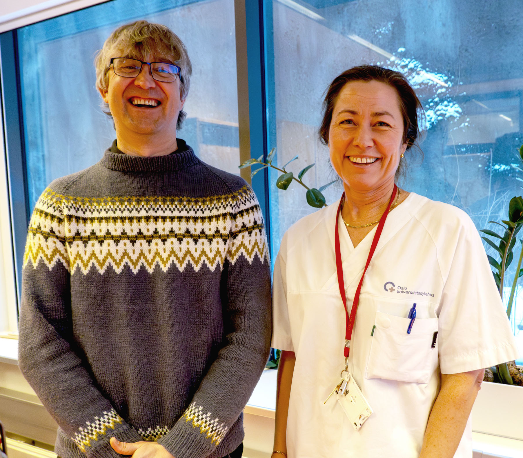 Mann og kvinne ser på kamera og smiler. Mann i ullgenser og med briller, kvinne i hvitt sykehusklær og med langt hår i hestehale.