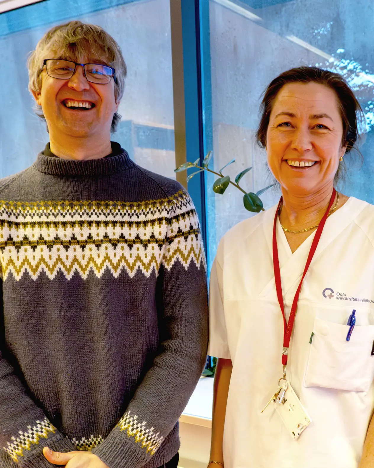 Mann og kvinne ser på kamera og smiler. Mann i ullgenser og med briller, kvinne i hvitt sykehusklær og med langt hår i hestehale.
