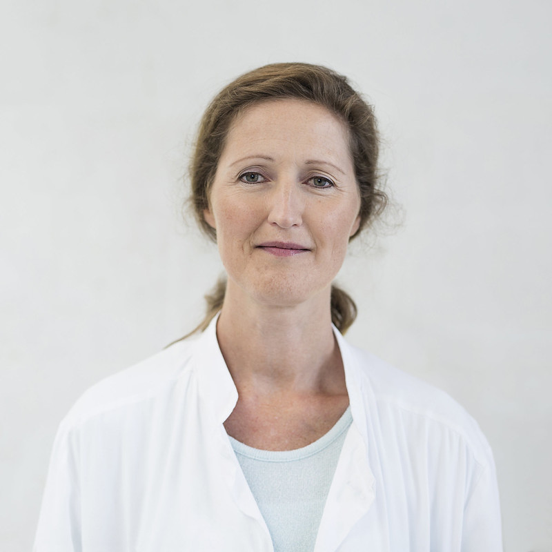 Kvinne med langt hår og i hvit sykehusfrakk, ser i kamera og smiler svakt. Foto.