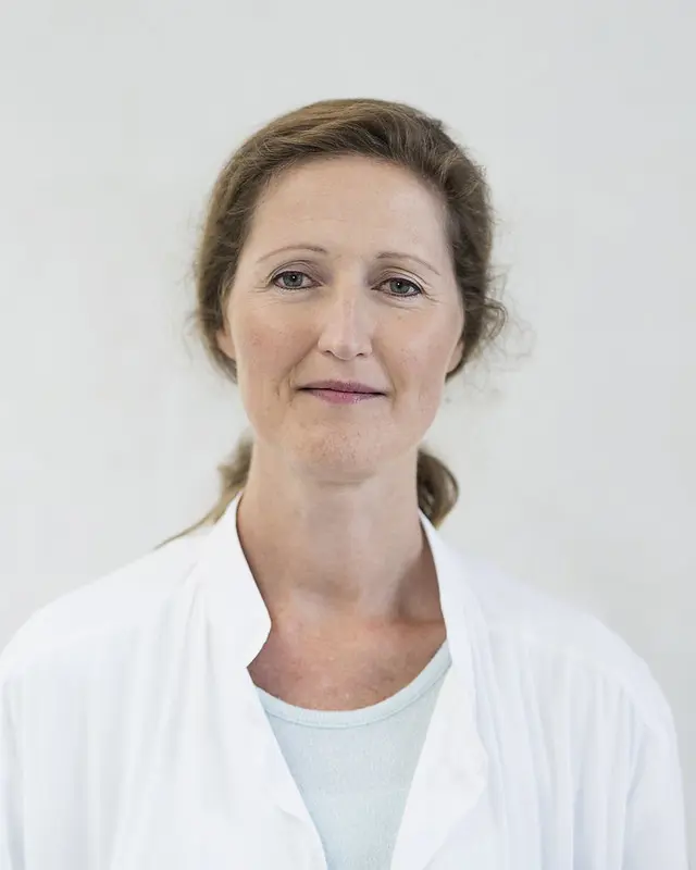 Kvinne med langt hår og i hvit sykehusfrakk, ser i kamera og smiler svakt. Foto.