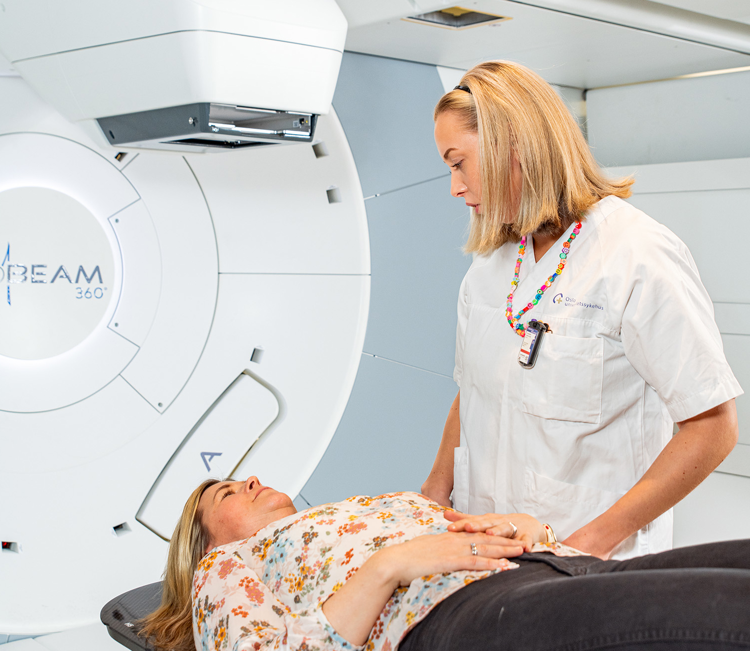 En kvinne ligger i en maskin på et sykehus i vanlige klær. Ved siden av står en sykepleier. Foto.