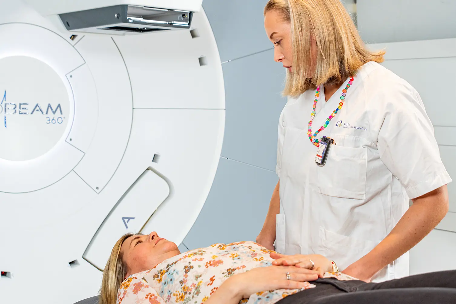 En kvinne ligger i en maskin på et sykehus i vanlige klær. Ved siden av står en sykepleier. Foto.