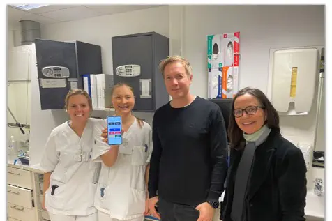En gruppe mennesker som poserer for et bilde. To i sykehusklær. Viser en skjerm på telefon.
