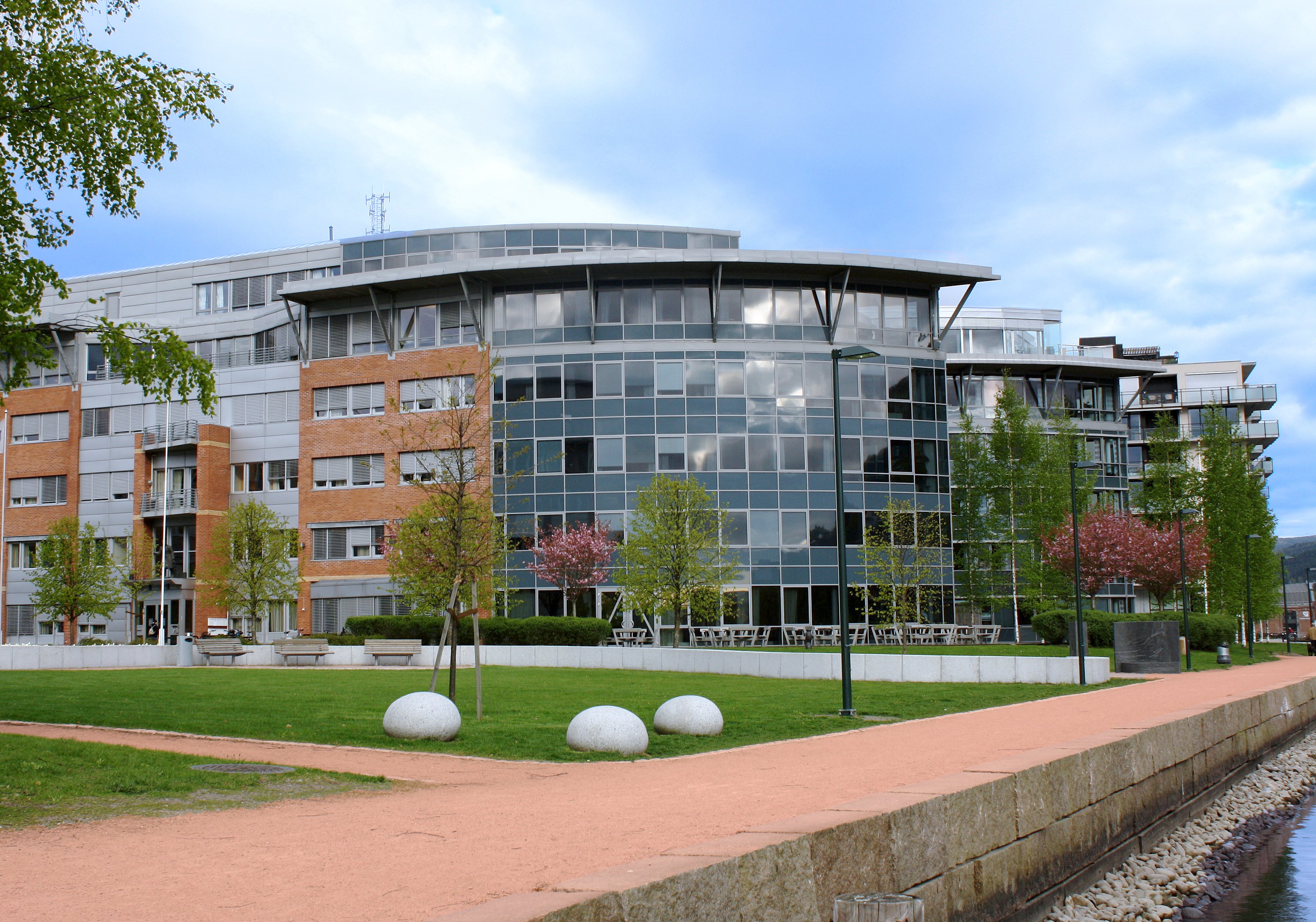 En bygning i murstein og glass med gangvei, trær og gress foran. Foto.