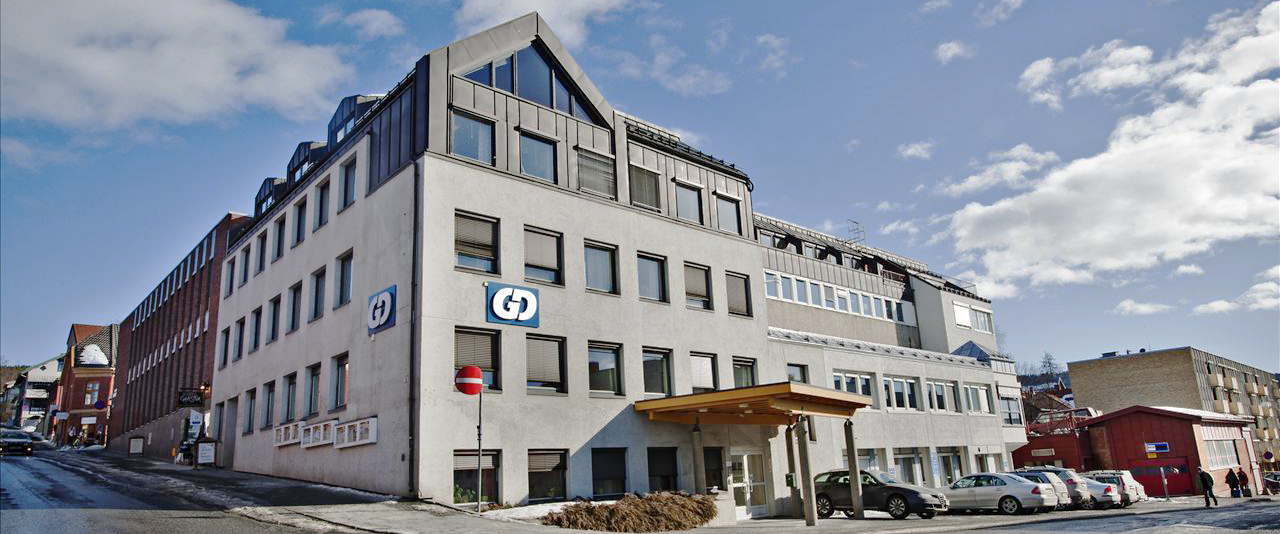 En kontorbygning i grå mur i en bygate med biler parkert foran. Foto.