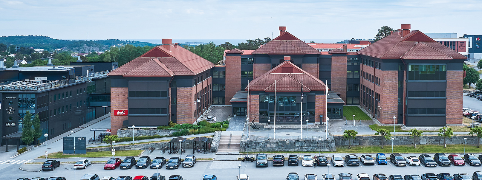 Stor, rød teglsteinsbygning i flere fløyer og med en stor parkeringsplass foran. Foto.