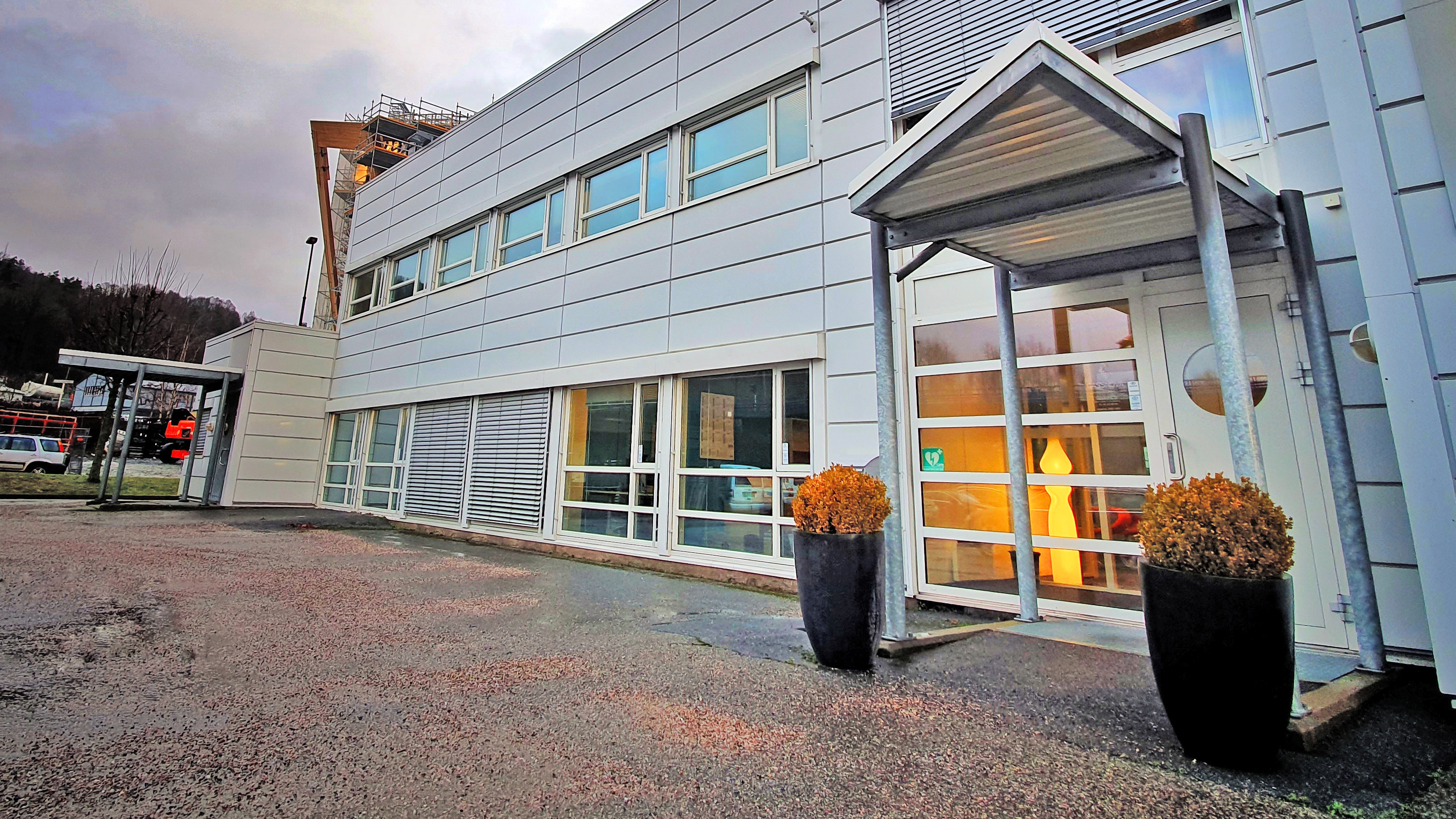 Inngangsparti på hvit kontorbygning med gruset parkeringsplass og blomster ved inngangsdør. Foto.