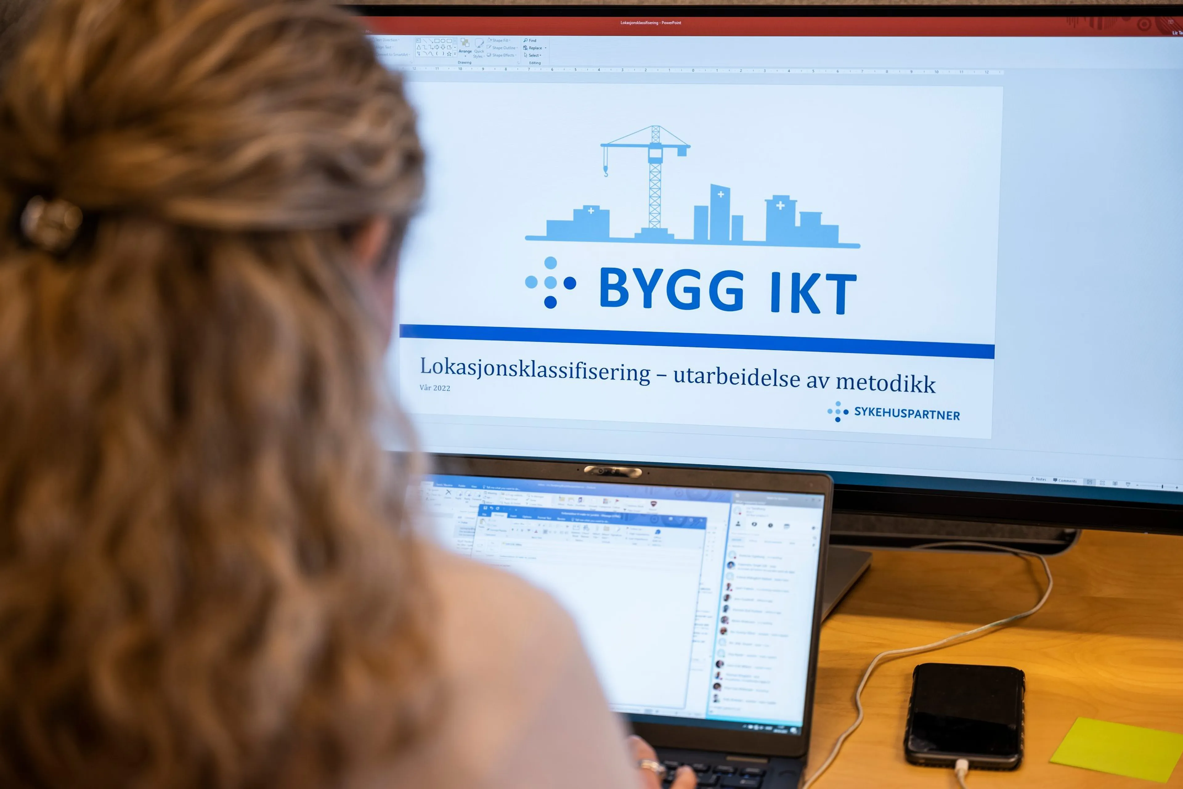 Bakholdet på kvinne som ser på to PC-skjermer. Set står "Bygg IKT" på den ene skjermen. Foto.