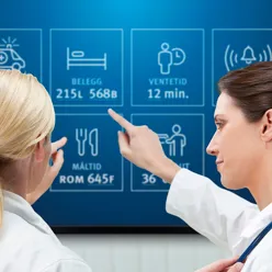 To helsepersonell foran skjerm som viser belegg, ventetid, måltid osv.