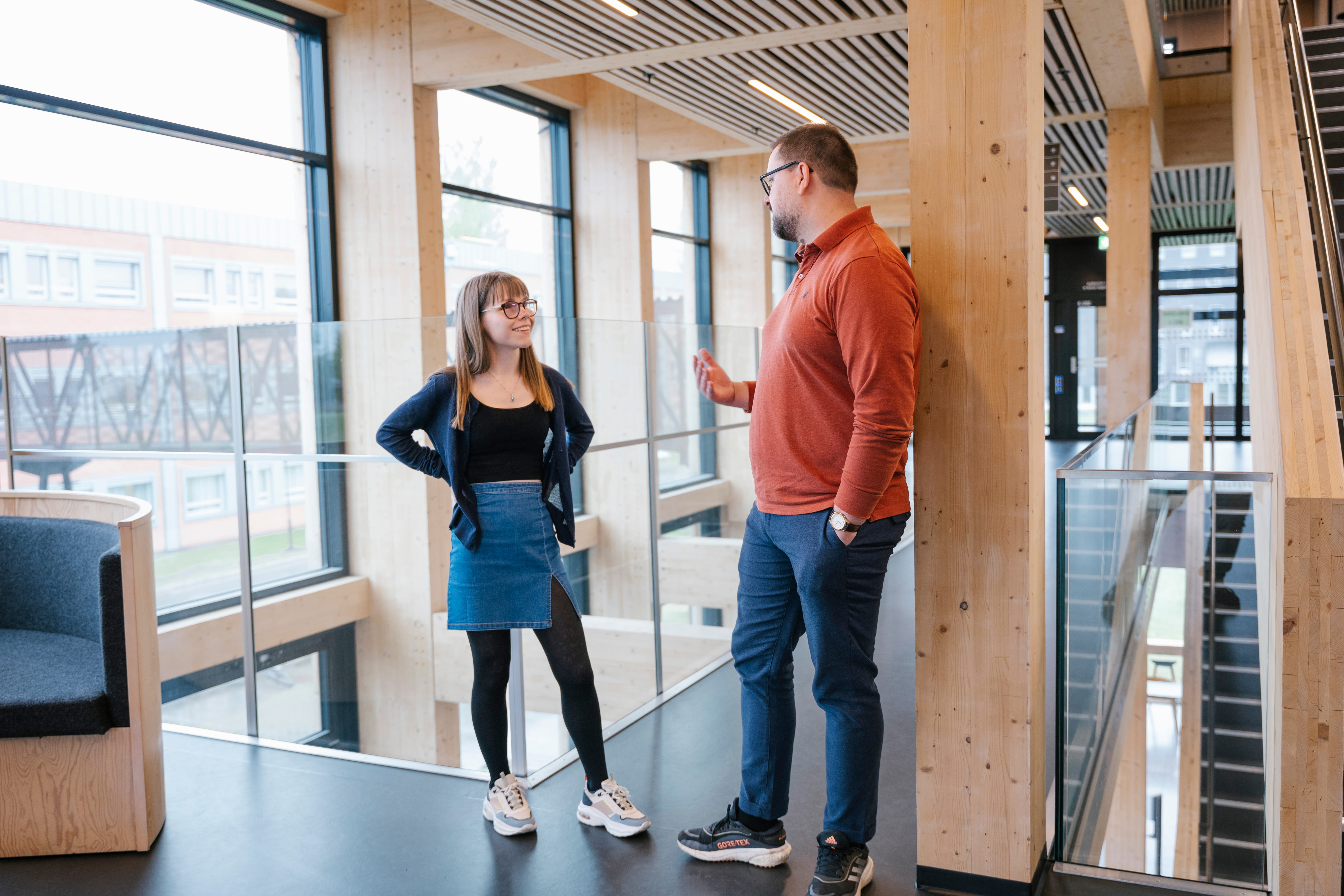 En mann og en kvinne snakker sammen i et åpent kontorlokale med store vindu og glassrekkverk. Foto.