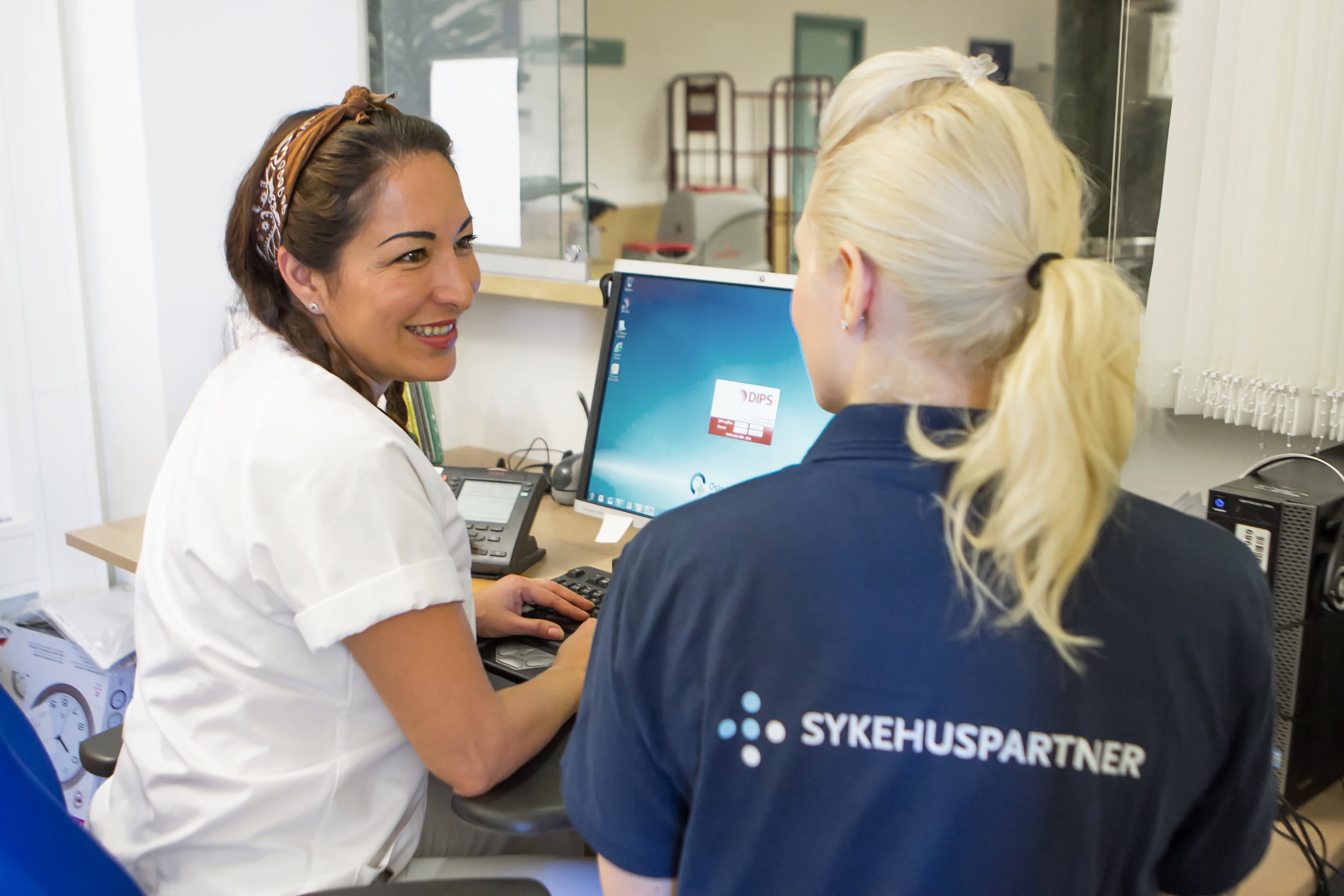 To kvinner snakker foran en PC, en i hvit sykepleier-skjorte, en i blå t-skjorte med påskriften "Sykehuspartner" på ryggen. Foto.