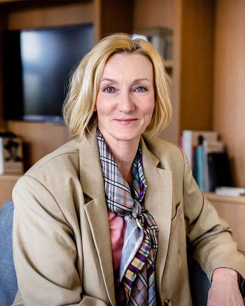 Smilende kvinne med blondt, halvlangt hår og beige dressjakke. Foto.