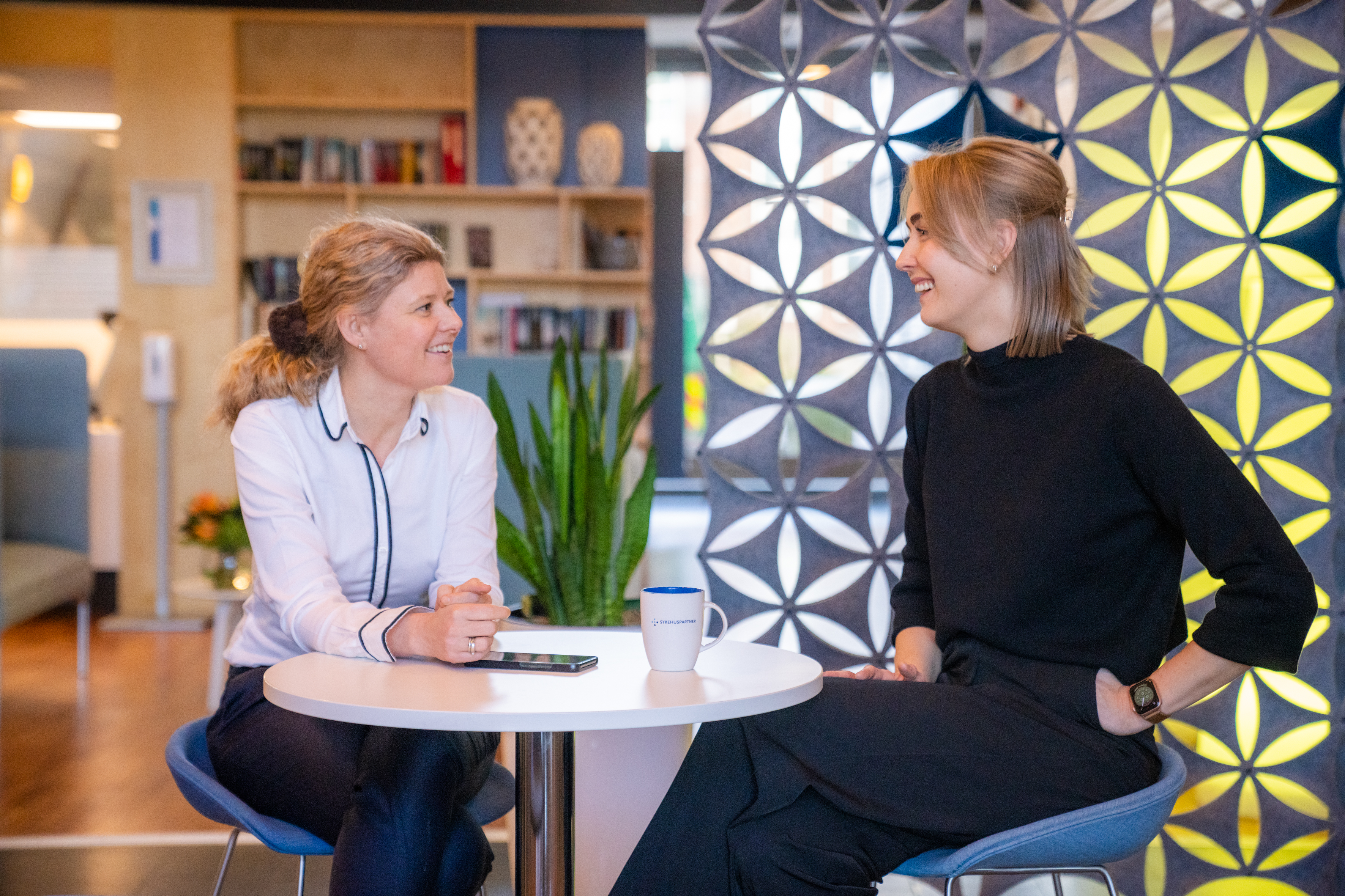 To damer som ser på hverandre og sitter ved et bord. Foto.