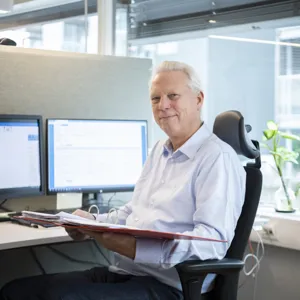 Eldre mann i kontorstol med perm i hendene og to pc-skjermer på pulten