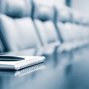 Skriveblokk på bord med mange stoler rundt. Foto.