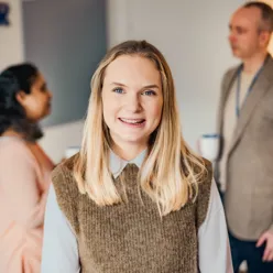 Smilende dame som ser i kamera og to personer som snakker bak henne. Foto.