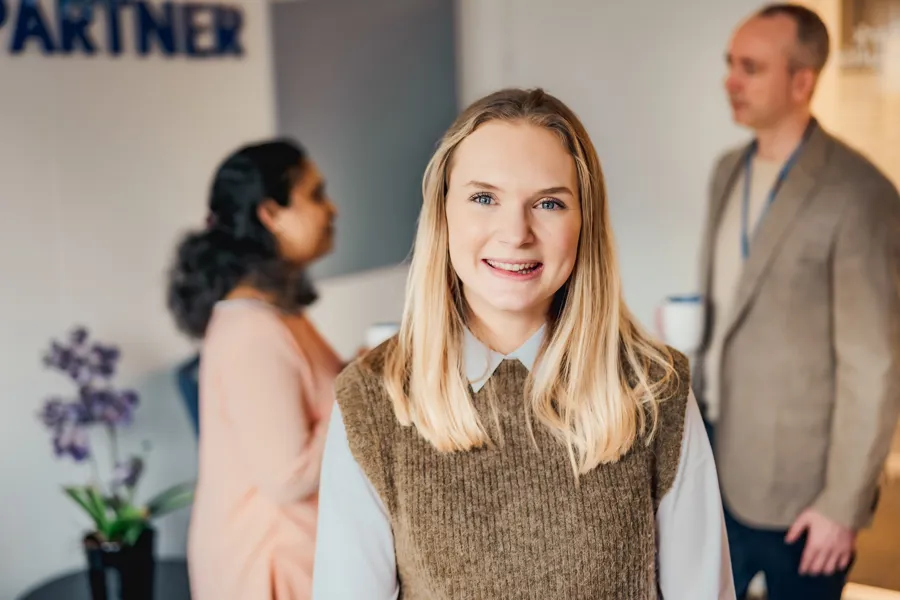 Smilende dame som ser i kamera og to personer som snakker bak henne. Foto.