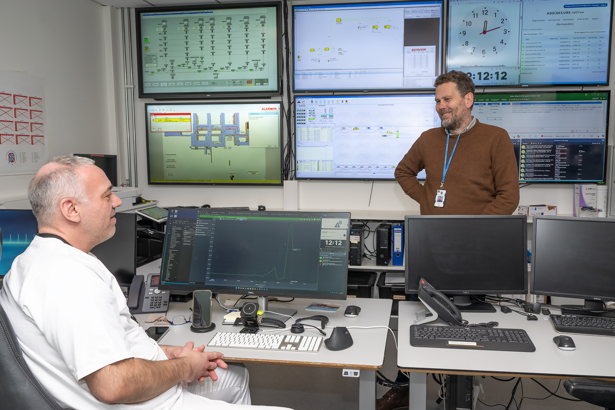 To menn i et rom med mange datamaskiner og mange dataskjermer på veggen. Foto.