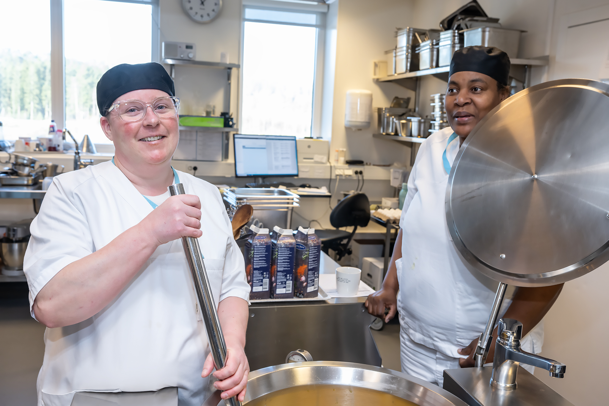 To smilende kvinner iført kokkeutstyr, på et industrikjøkken. Foto.