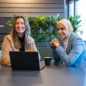 To kvinner sitter ved et bord med en pc mellom seg. En med langt, blondt hår, en med hijab. Foto.