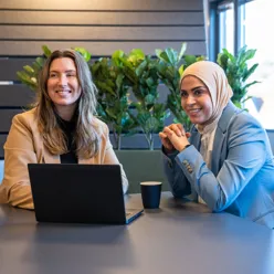 To kvinner sitter ved et bord med en pc mellom seg. En med langt, blondt hår, en med hijab. Foto.
