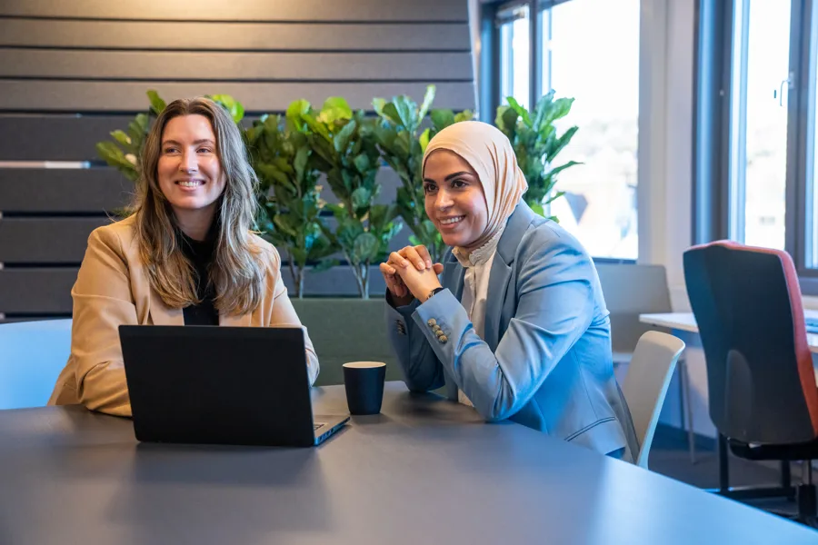 To kvinner sitter ved et bord med en pc mellom seg. En med langt, blondt hår, en med hijab. Foto.