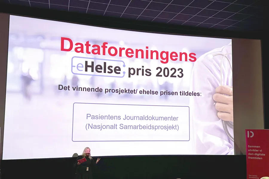 Skjerm på scene med teksten "Dataforeningens eHelsepris 2023. Det vinnende prosjektet tildeles: Pasientens journaldokumenter. Nasjonalt samarbeidsprosjekt". Foto.