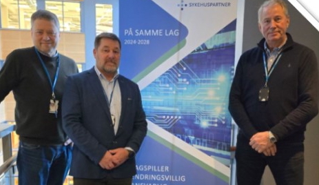 Tre menn som står foran en plakat med Sykehuspartners slagord, På samme lag. Foto.