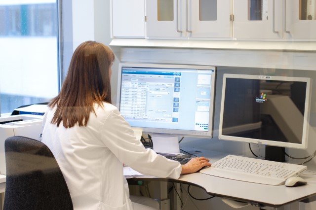 Kvinne i hvit sykehusfrakk sitter med ryggen til, vendt mot en pc på et kontor. Foto.