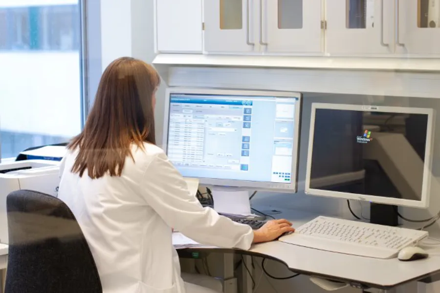 Kvinne i hvit sykehusfrakk sitter med ryggen til, vendt mot en pc på et kontor. Foto.