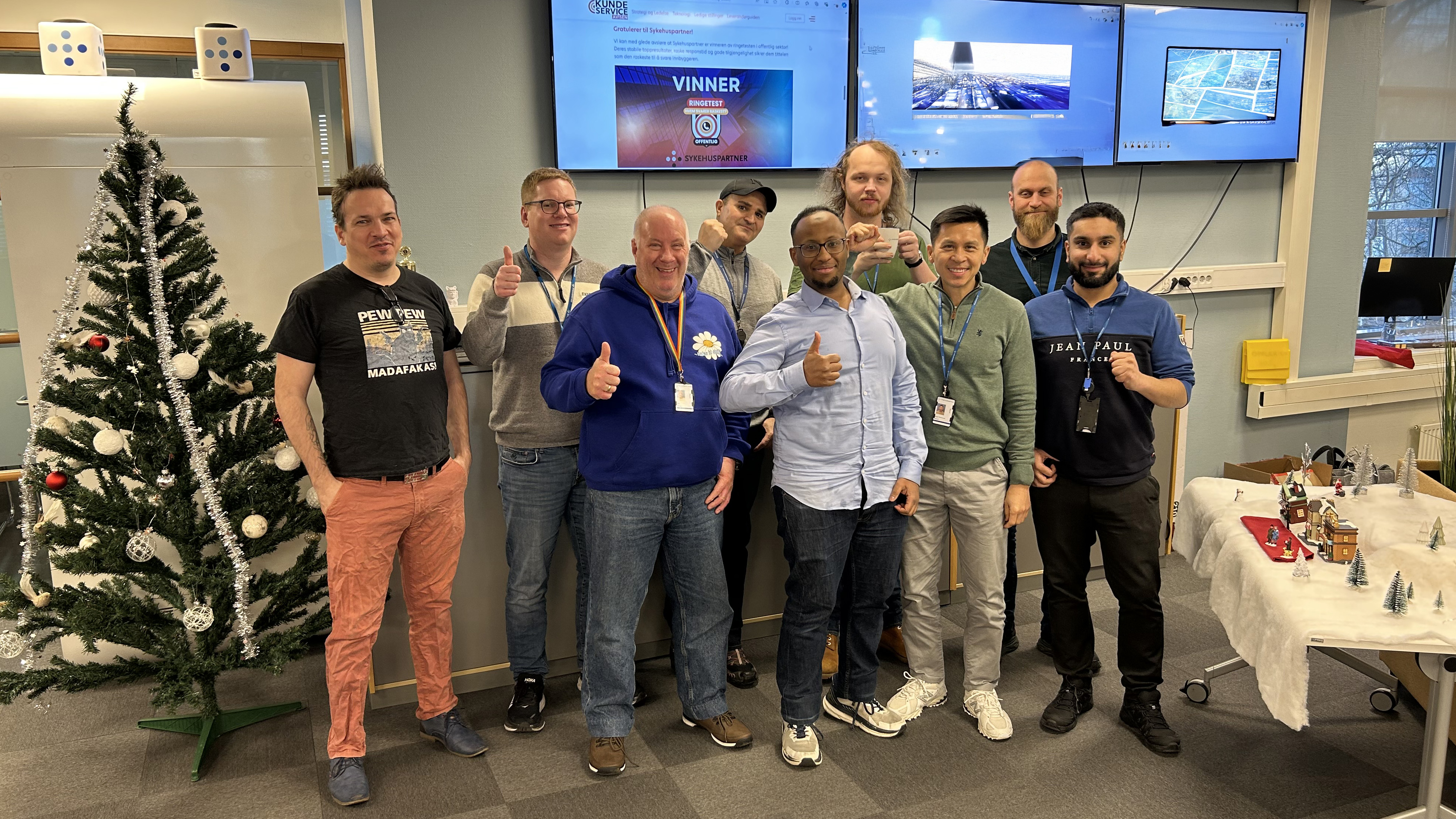 En gruppe mennesker i kontormiljø som poserer for et bilde, noen viser tommel opp. Foto.