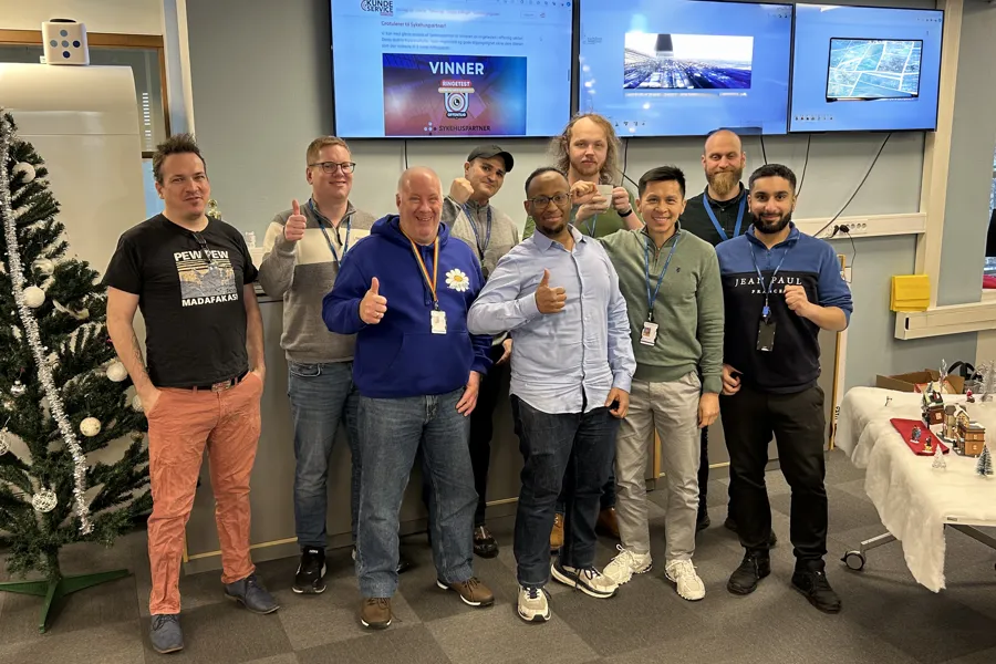 En gruppe mennesker i kontormiljø som poserer for et bilde, noen viser tommel opp. Foto.