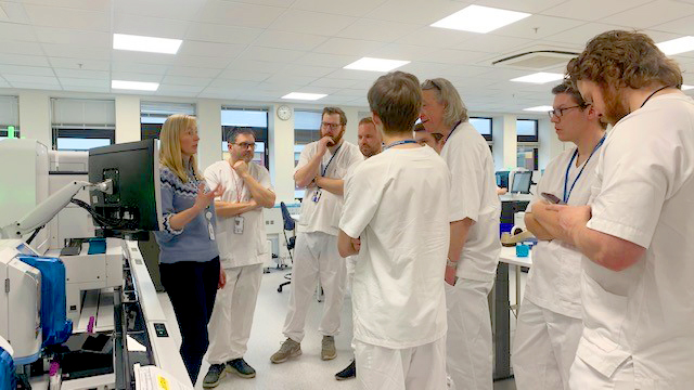 En gruppe menn i hvite sykehusklær lytter til en kvinne i blå genser, inne på et kontor med datautstyr.  Foto.