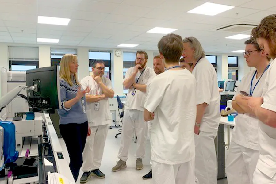 En gruppe menn i hvite sykehusklær lytter til en kvinne i blå genser, inne på et kontor med datautstyr. Foto.