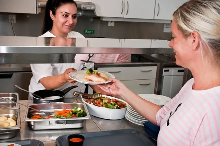 Kantinepersonal gir tallerken med mat til en kunde over en matdisk i kantinemiljø. Foto.