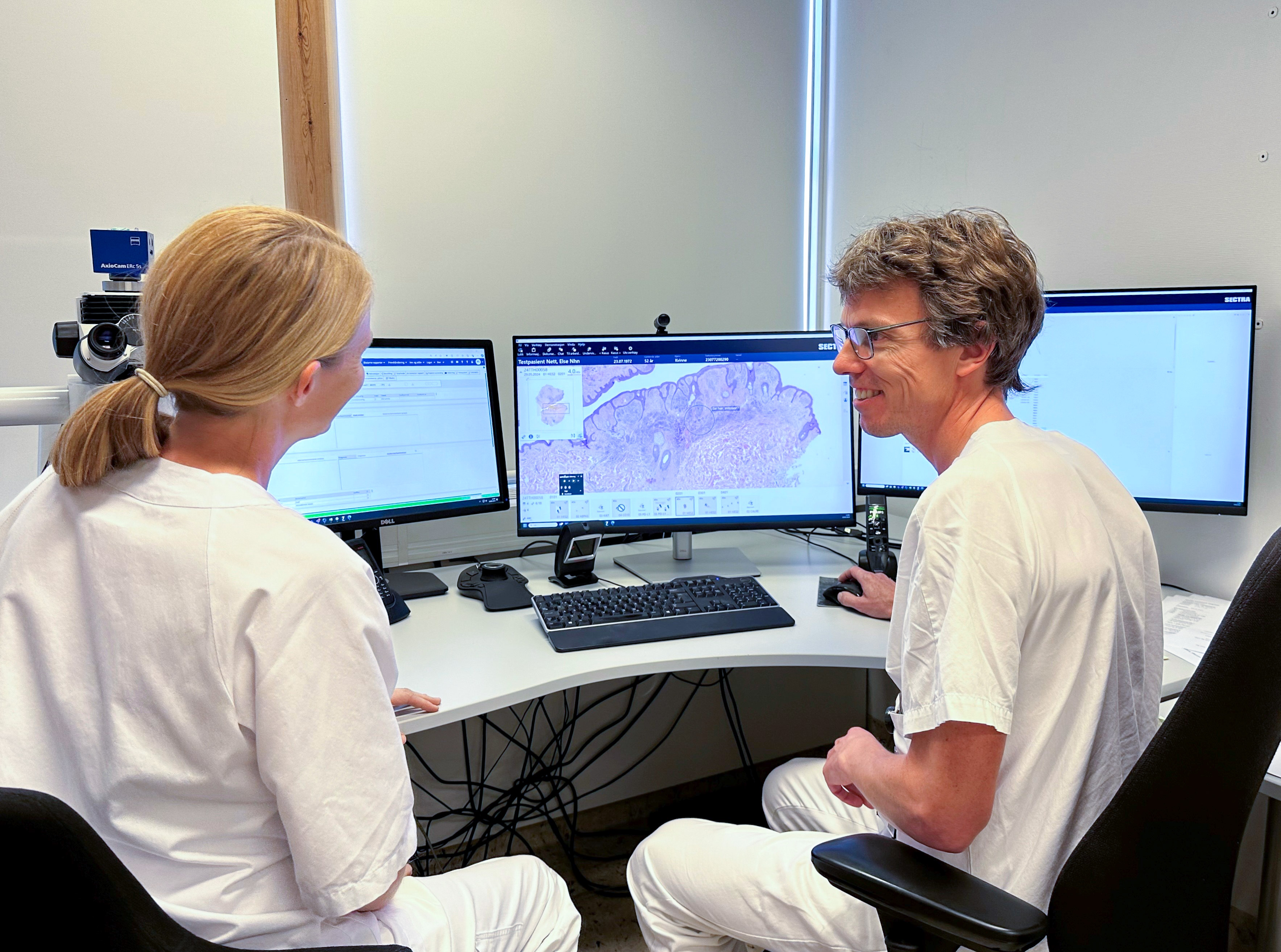 Mann og dame i hvitt sykehustøy sitter ved en pult med dataskjermer. Foto.