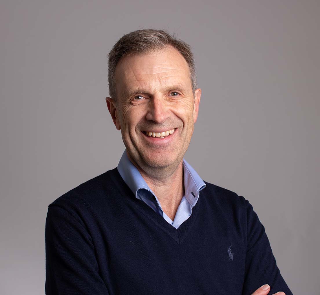 Smilende mann med grått hår, skjorte og genser. Foto.