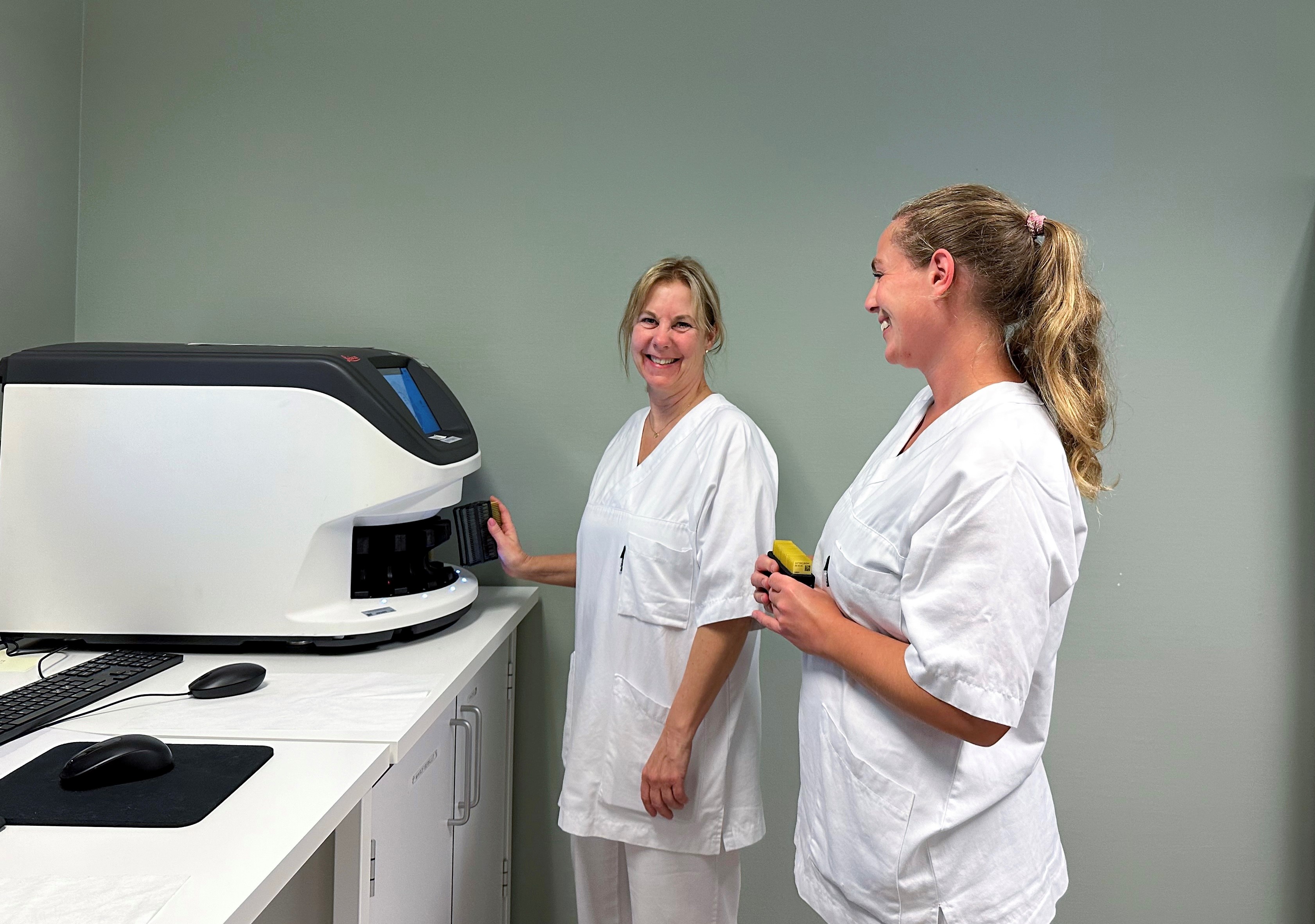 To smilende kvinner i hvitt sykehustøy står ved en maskin. Foto.