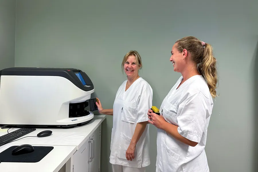 To smilende kvinner i hvitt sykehustøy står ved en maskin. Foto.