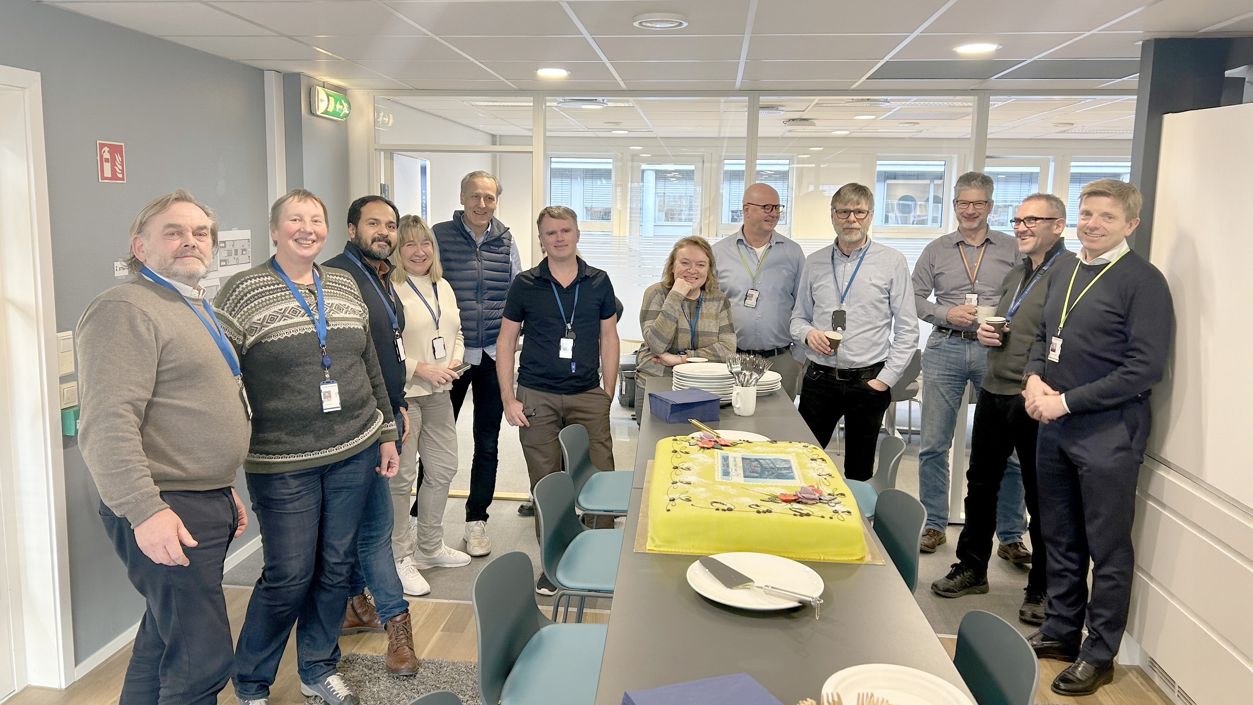 En gruppe mennesker står rundt et bord på et møterom. På bordet står en stor, gul kake. Foto.