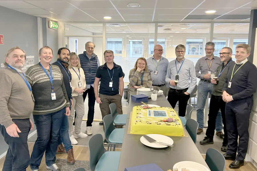 En gruppe mennesker står rundt et bord på et møterom. På bordet står en stor, gul kake. Foto.