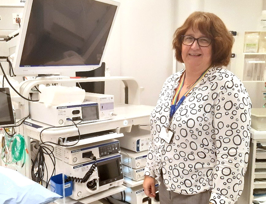 Smilende kvinne som står ved siden av mange maskiner, ved en sykehusseng. Foto.