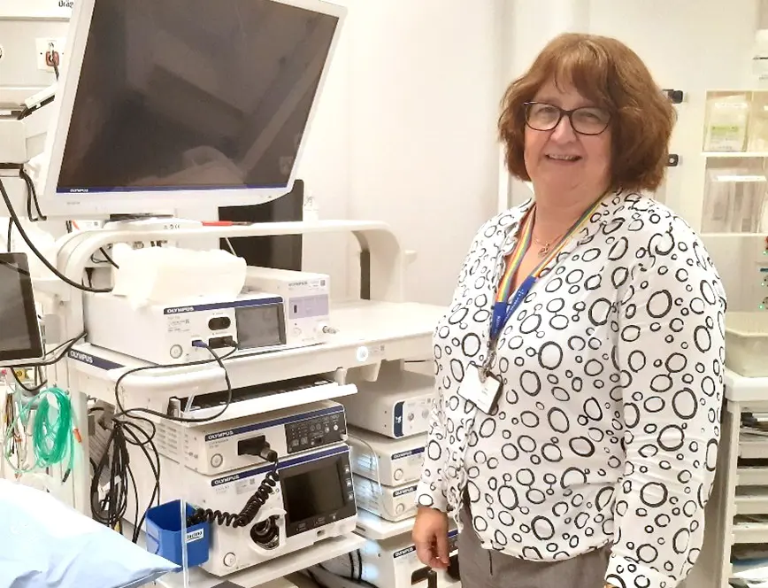 Smilende kvinne som står ved siden av mange maskiner, ved en sykehusseng. Foto.