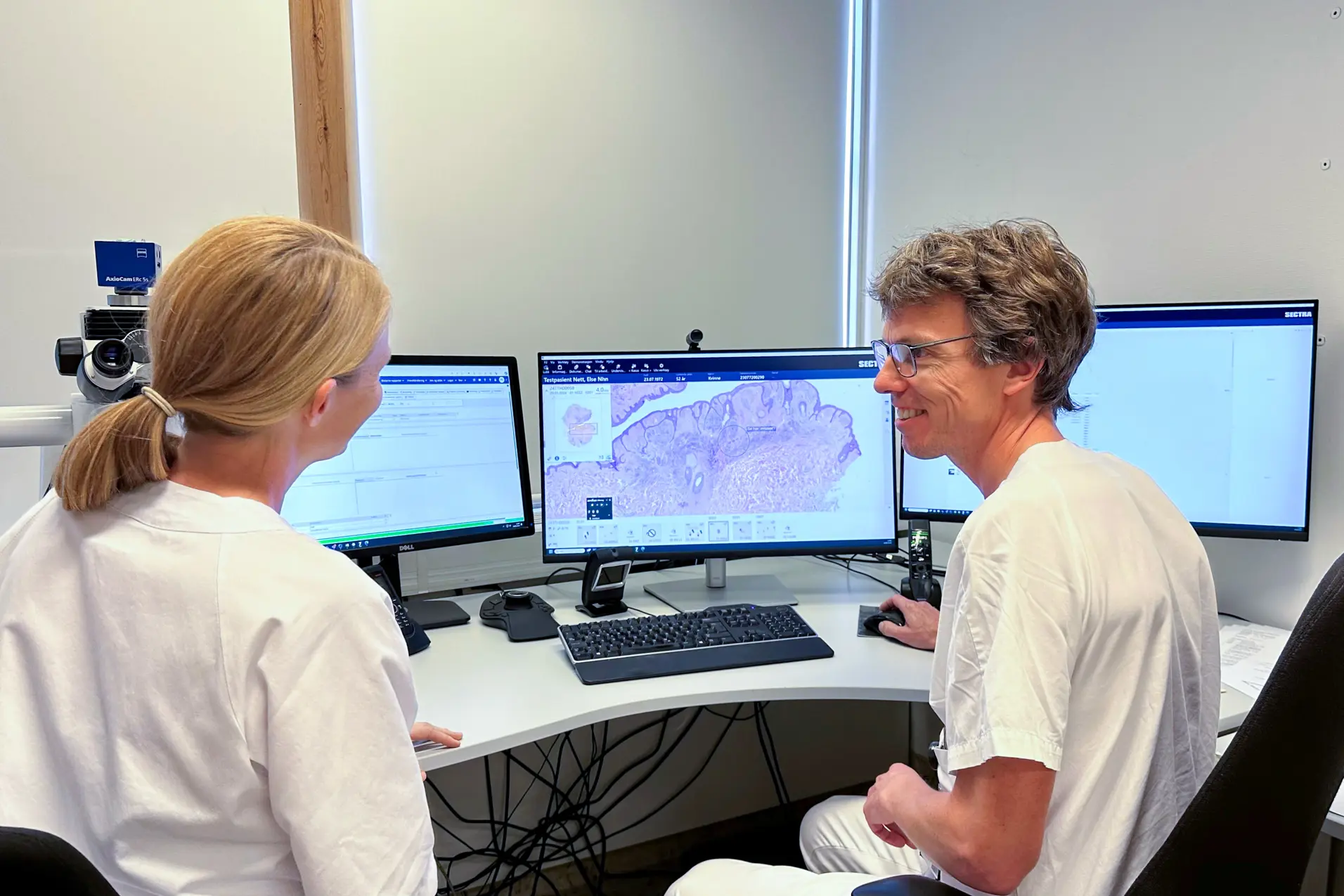 En mann og en kvinne med sykehusklær som sitter ved et skrivebord med datamaskiner. Foto.