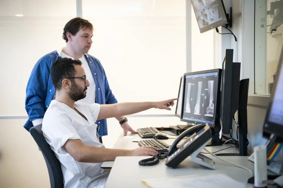 To menn i syhehusklær ser på en dataskjerm. Foto.