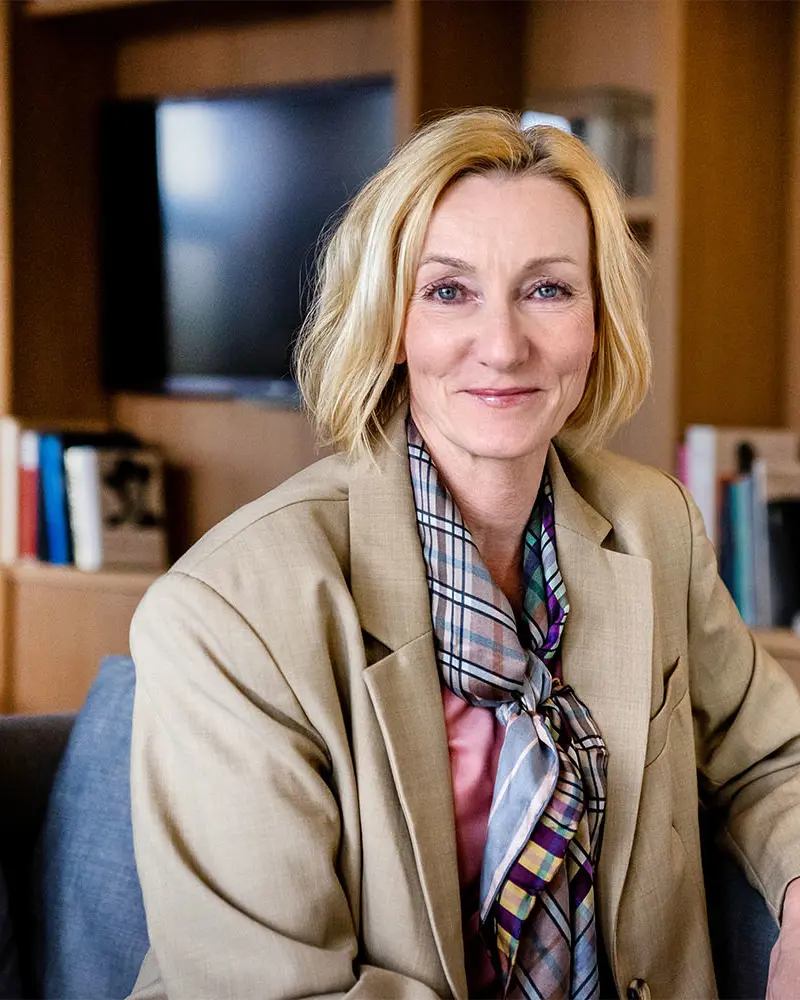 Smilende kvinne med blondt, halvlangt hår og beige dressjakke.